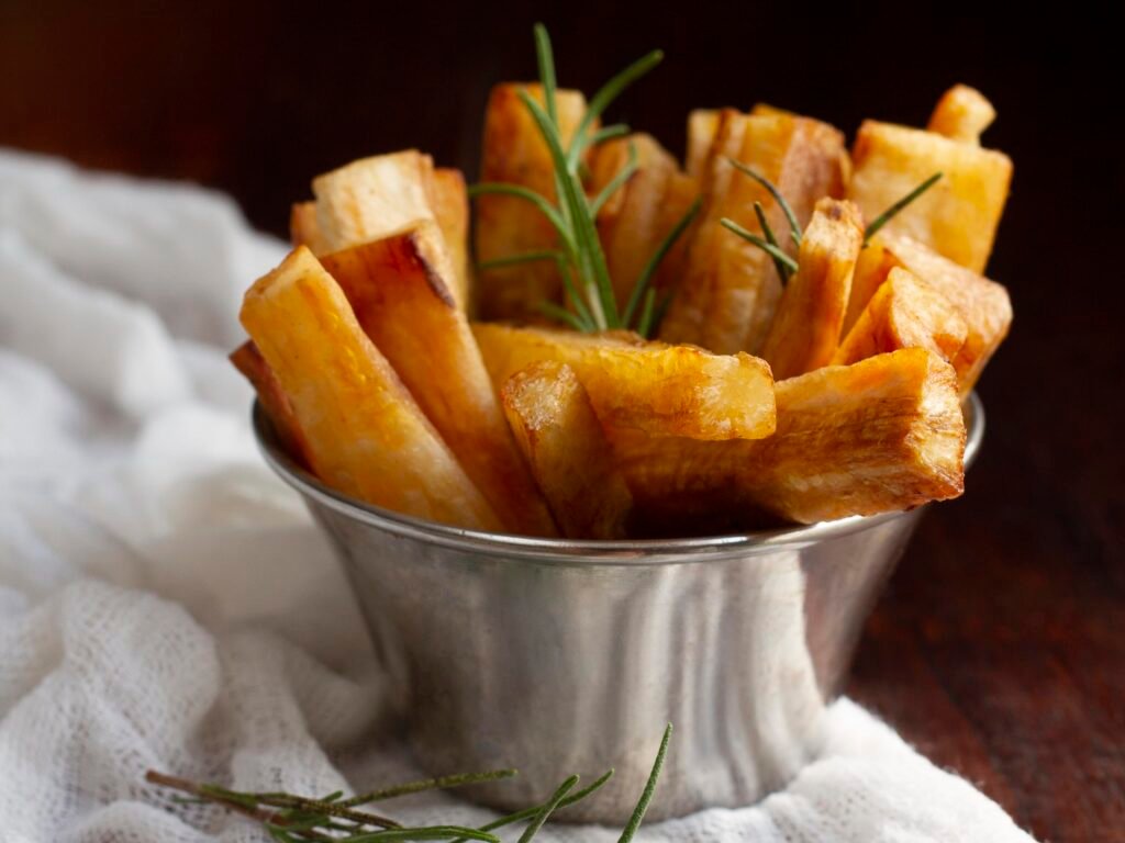 Uma tigela de metal prateada cheia de palitos fritos e dourados de mandioca, com um raminho de alecrim no topo, está sobre um pano de prato branco amassado, que repousa sobre uma superfície de madeira escura. Há um segundo raminho de alecrim na frente da tigela. O fundo é escuro e desfocado.