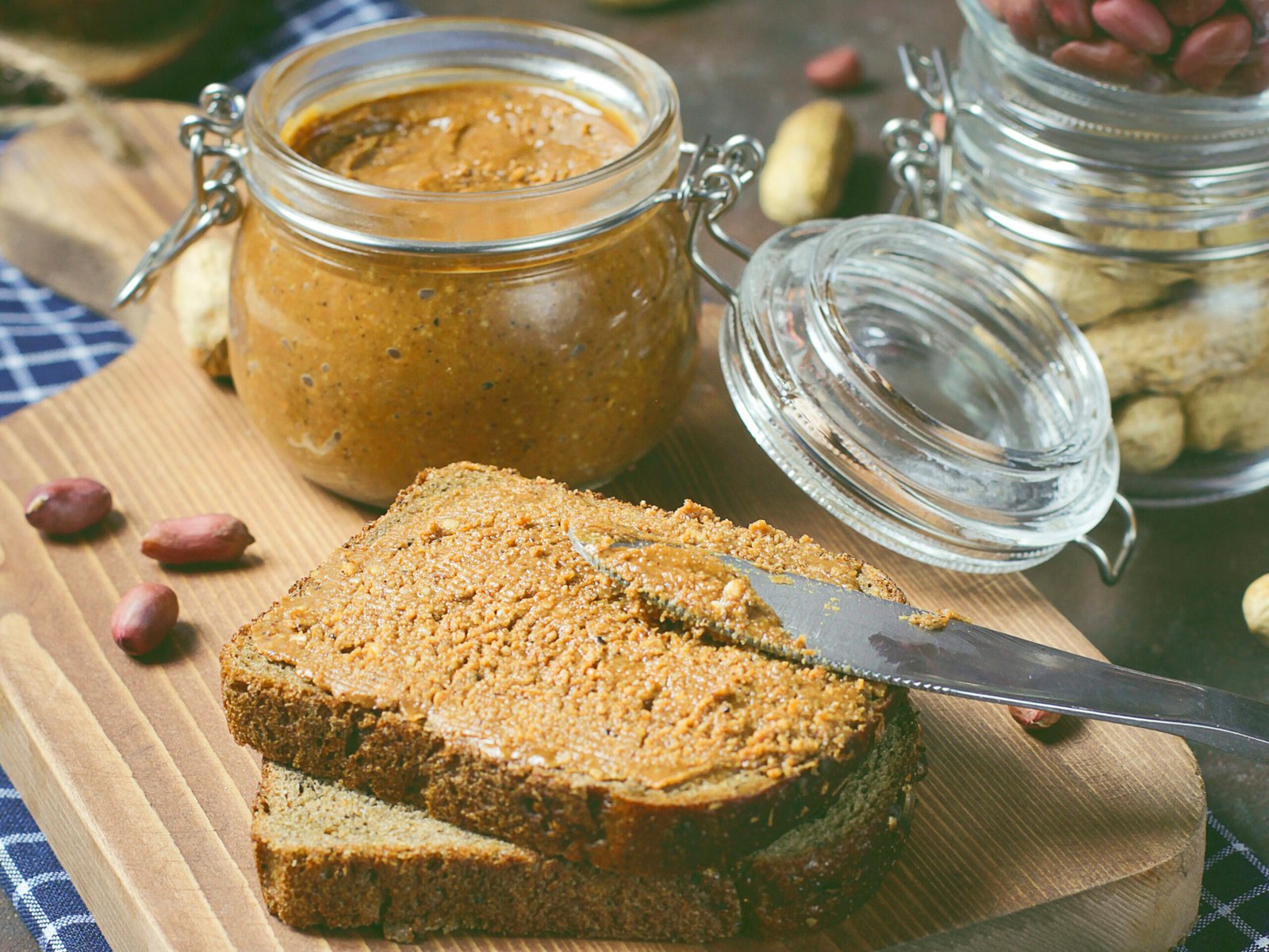 Duas fatias de pão integral com manteiga de amendoim espalhada nelas estão sobre uma tábua de madeira clara. Uma faca está sobre a fatia superior, e um pote aberto de manteiga de amendoim está atrás do pão. Um frasco com amendoins dentro está à direita, e amendoins soltos estão espalhados pela superfície escura da mesa. Um pano de prato azul e branco está sob a tábua de madeira.