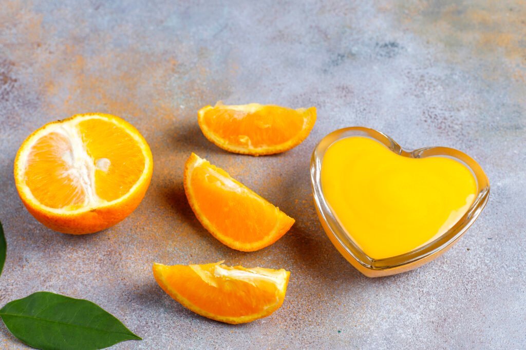 Laranja cortada no meio com algumas fatias ao lado e um pote de vidro em formato de coração cheio de suco de laranja