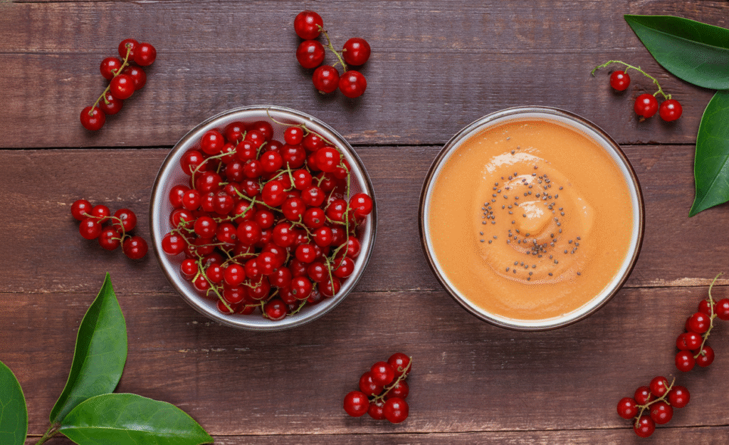 Uma composição vista de cima mostra dois bowls sobre uma superfície de madeira escura. À esquerda, um bowl contém um monte de acerolas frescas e vermelhas. À direita, um bowl com mousse vegano de acerola e chia, de cor laranja clara e translúcida, com sementes de chia espalhadas no topo. Várias acerolas extras e algumas folhas verdes estão espalhadas pela superfície de madeira, criando um ambiente natural e convidativo.