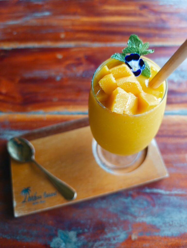 Copo de suco de manga haden e abacaxi com pedaços de manga em cima e uma flor e hortelã para decorar em cima de uma tabua de madeira e uma mesa de madeira