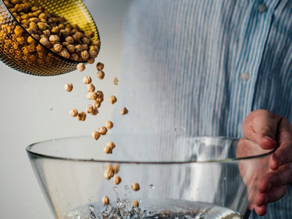 Pessoa derrubando uma vasilha de vidro marrom de grão de bico em outra vasilha de vidro trasnparente cmo água.