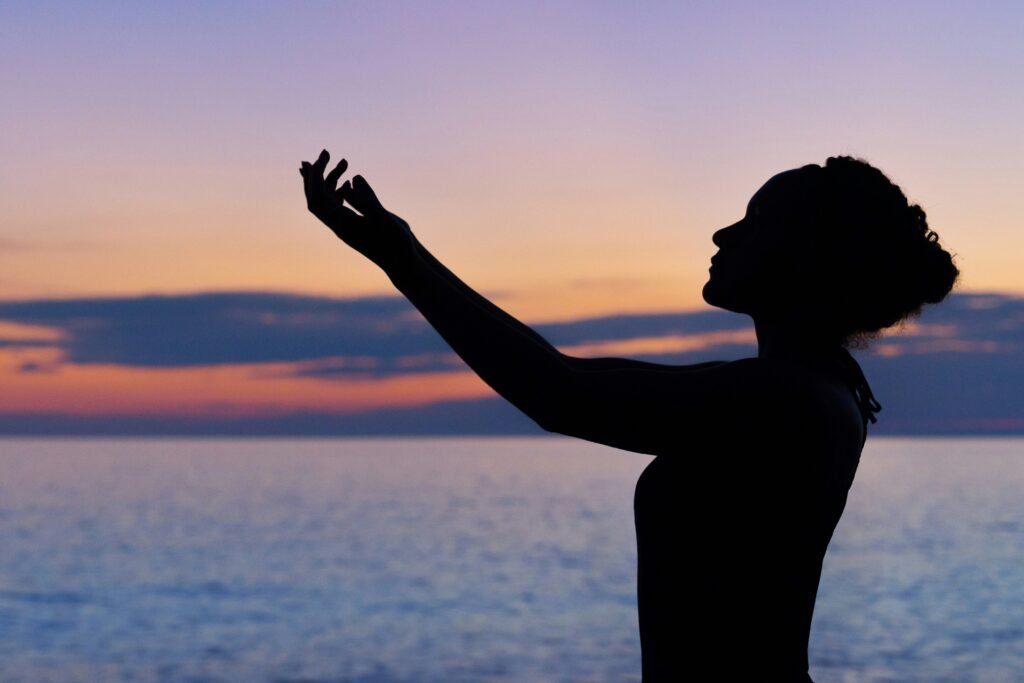Silhueta de uma mulher com as mãos para cima praticando mindfulness em frente ao mar em um pôr do sol